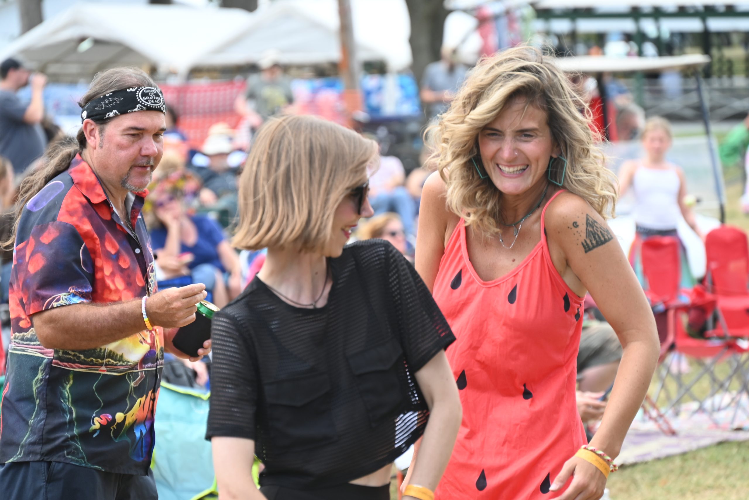 Dancing Patrons – photo by Kevin Slick Two young women, one wearing a watermelon-themed dress and the other in a black t-shirt, and a man with a bright show shirt smile and dance together at the 2025 Watermelon Pickers' Fest