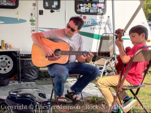 Jack and JJ playing tunes – Photo by Chester SImpson Jack Dunlap and his son JJ seated at a campsite, plays guitar with his young son, JJ, who is playing mandolin.