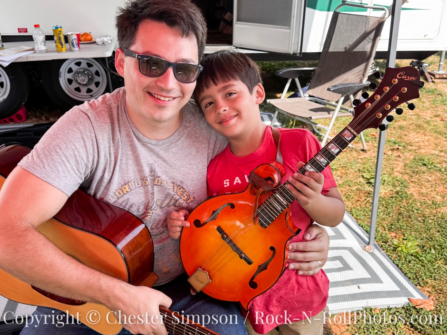 Jack Dunlap and his son JJ sit together and smile for a photo. Jack is holidng his guitar and JJ has a Carey mandolin